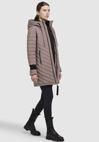 Polstret parkajakke i taupe-farve med hætte, skråt quiltet design og sorte detaljer. Har frontlynlås og sidelommer. Bæres over sorte leggings og støvler.