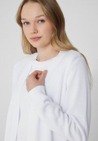 Cardigan en tricot blanc avec un avant ouvert, des poignets côtelés et une texture douce. Le modèle tient doucement le col du cardigan, créant une apparence décontractée.