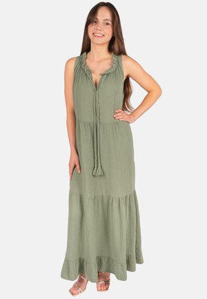Frau steht mit der Hand auf der Hüfte und trägt ein ärmelloses olivgrünes Maxikleid mit Rüschen am Ausschnitt und Quastenbändern, sie lächelt.