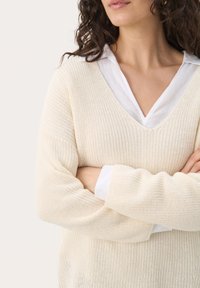 Knit cream sweater with a v-neck, featuring a textured pattern. Worn over a white collared shirt, arms crossed, showcasing relaxed sleeves.