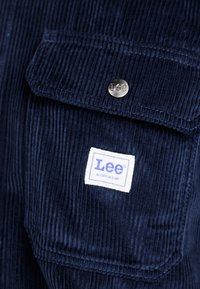 Tumesinine corduroy tööjakk, millel on suur rinnatasku, metallne nööbiklipp ja valge silt violetse tekstiga.