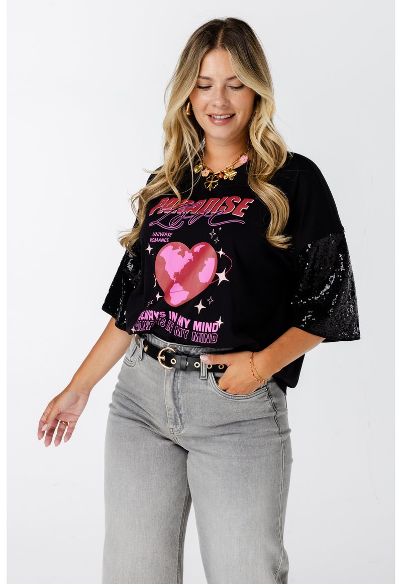 Zwarte t-shirt met roze grafische print met een hart en tekst. Korte pailletten mouwen. Gecombineerd met lichtgrijze jeans en een zwarte riem.