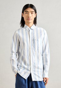 Chemise boutonnée rayée bleu clair et blanche avec un col, une poche poitrine et des manches longues, fabriquée à partir d'un tissu texturé.