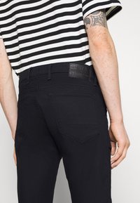 Pantalones de mezclilla negros con una etiqueta de G-Star en la cinturilla, con un corte ajustado y dos bolsillos traseros con diseños angulares. Camisa de rayas parcialmente visible.