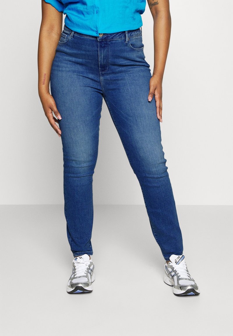 Persona vistiendo jeans azules ajustados, zapatillas blancas y grises, y una camisa de botones azul brillante, de pie contra un fondo sencillo.