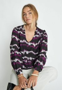 Blusa nera con motivo a zigzag viola e bianco, scollatura a V e maniche lunghe a sbuffo. Indossata con pantaloni chiari e un accessorio nero.
