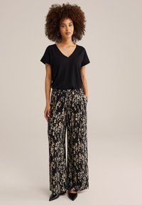 T-shirt noir à col en V associé à un pantalon évasé avec un motif abstrait noir et beige. Tissu texturé et poches latérales visibles.