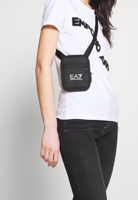 Czarna torba crossbody wykonana z trwałego materiału, wyposażona w zapinaną na zamek przednią kieszeń oraz białe logo EA7. Regulowany pasek dla wygody noszenia.