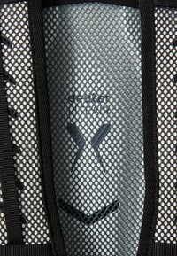 Schwarzer Mesh-Rucksack mit einem gemusterten Außenmaterial, verstärkten Kanten und einem Logo-Design in Dunkelgrau. Atmungsaktives Material für Belüftung.