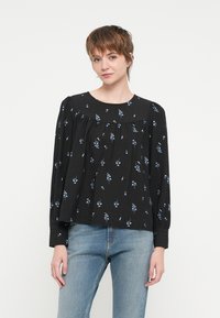 Svart bluse med lange ermer i lett stoff, prydet med et blomsterdesign i blått. Rynkete detaljer ved brystet skaper en luftig silhuett.