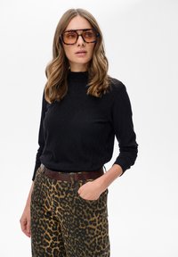 Kobieta w dużych okularach w tortoishell, czarnej, fakturowanej bluzce z długim rękawem i spodniami w panterkę z brązowym paskiem, z ręką w kieszeni.