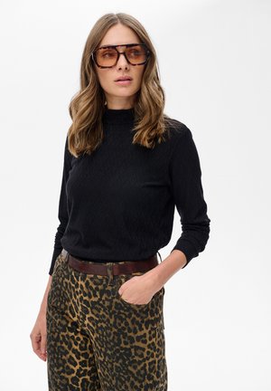 Vrouw met grote schildpadvormige zonnebril, een zwart textuur langemouwen top en een broek met luipaardprint met een bruine riem, hand in de zak.