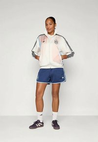 Λευκό αθλητικό μπουφάν με παστέλ λεπτομέρειες, μαύρες ρίγες, σε συνδυασμό με ναυτικά σορτς και μαύρα αθλητικά παπούτσια. Περιέχει λογότυπο στο μπουφάν και στα σορτς.