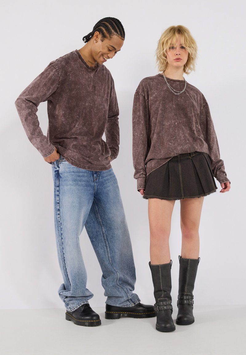 To modeller står side om side. Den ene har på seg en falmet brun langermet skjorte og lyseblå jeans. Den andre har på seg en brun hettegenser, svart miniskjørt og svarte knelengde støvler.