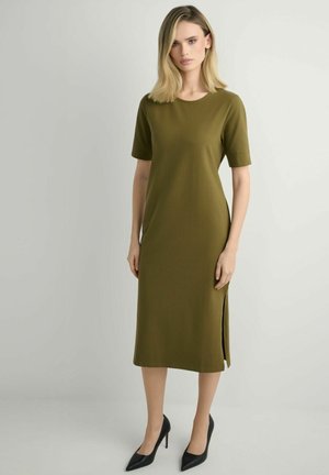 Kobieta w oliwkowozielonej, sięgającej kolan sukience z krótkim rękawem i rozcięciem po boku, stojąca na czarnych szpilkach na jednolitym tle.