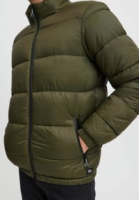 Grønn pufferjakke med høy krage, glidelås og sidelommer med svarte detaljer. Tekstured, quiltet design for isolasjon.