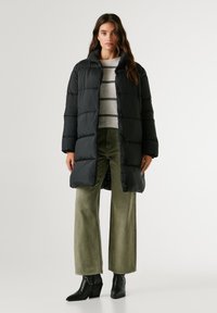 Melna pildīta mētelis ar augstu apkakli, ar horizontālām šuvēm. Valkāts virs gaiši svītrainā džempera un oliņzaļām platām biksēm.