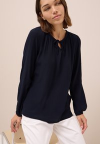 Blusa blu navy con maniche lunghe, scollatura arricciata e dettaglio con fiocco. Realizzata in tessuto morbido, abbinata a pantaloni bianchi.