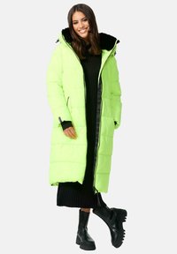 Neon zelena puffer jakna s kapuljačom obloženom crnom umjetnom krznom, zatvaranjem na rajsferšlus i bočnim džepovima, nosi se preko crne haljine s rebrastim uzorkom i crnih čizama.
