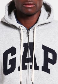 Felpa grigia con zip realizzata in mix di cotone, con logo "GAP" blu navy sul davanti, dettagli in tessuto scuro testurizzato e coulisse.