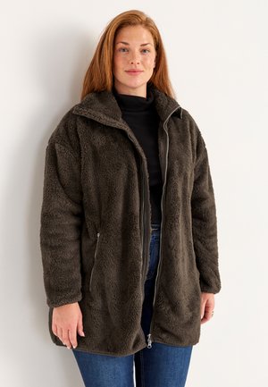 Flauschiger, olivgrüner Mantel aus weichem, strukturiertem Stoff. Mit einem Reißverschluss vorne, hohem Kragen und Eingrifftaschen. Kombiniert mit einem schwarzen Oberteil und blauen Jeans.