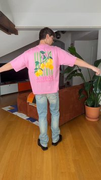 Mann som har på seg en rosa Adidas Originals t-skjorte med gul fruktdesign, lyseblå jeans og svarte sko, stående i en stue med planter.