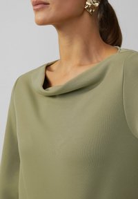 Haut vert olive avec un design à col drapé, texture lisse, manches longues et éclat subtil. Présente une boucle d'oreille dorée remarquable.