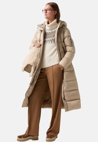 Vrouw in een beige lange pufferjas, witte gebreide coltrui, bruine broek en suède wandelschoenen, met een crèmekleurige bonten tas.