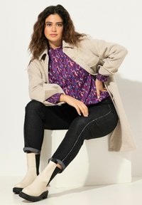 Abrigo de lana beige sobre una blusa morada con estampado, combinado con jeans negros con costuras blancas y botas de tobillo crema, sentado contra un fondo blanco.