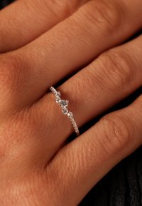 Zilveren ring met drie heldere edelstenen in een golvend ontwerp, met kleine accentstenen langs de band, gepresenteerd op een hand.