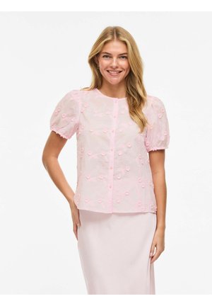 Femme souriante aux longs cheveux blonds portant un chemisier rose clair à manches bouffantes et broderies florales, associé à une jupe rose pâle.