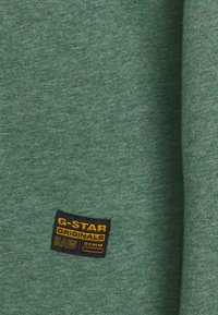 Grüner Stoff mit strukturiertem Strickmuster. Enthält einen schwarz-gelben G-Star-Logoaufnäher mit den Schriftzügen "ORIGINALS" und "RAW DENIM".