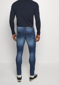 Jeans de denim azul ajustados con un lavado desgastado, detalles desgastados y una etiqueta negra en la cintura. Llevados con una camisa de manga larga azul marino y zapatillas.