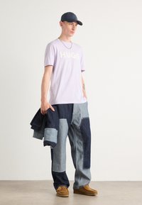 Light purple T-shirt with "HUGO" in white, paired with patchwork denim pants featuring various blue shades. Brown slip-on shoes. Holding a jacket.