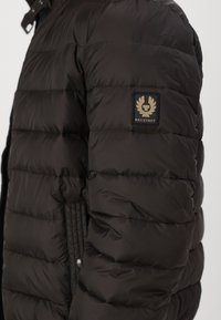 Tmavohnedá prešívaná bunda s kockovanou textúrou, s čiernou a zlatou nášivkou loga Belstaff na ľavom pleci a zapínaním na patentky.