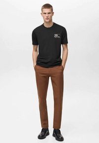 T-shirt nera a maniche corte con logo stampato sul petto, abbinata a pantaloni marroni slim-fit e scarpe nere stringate.