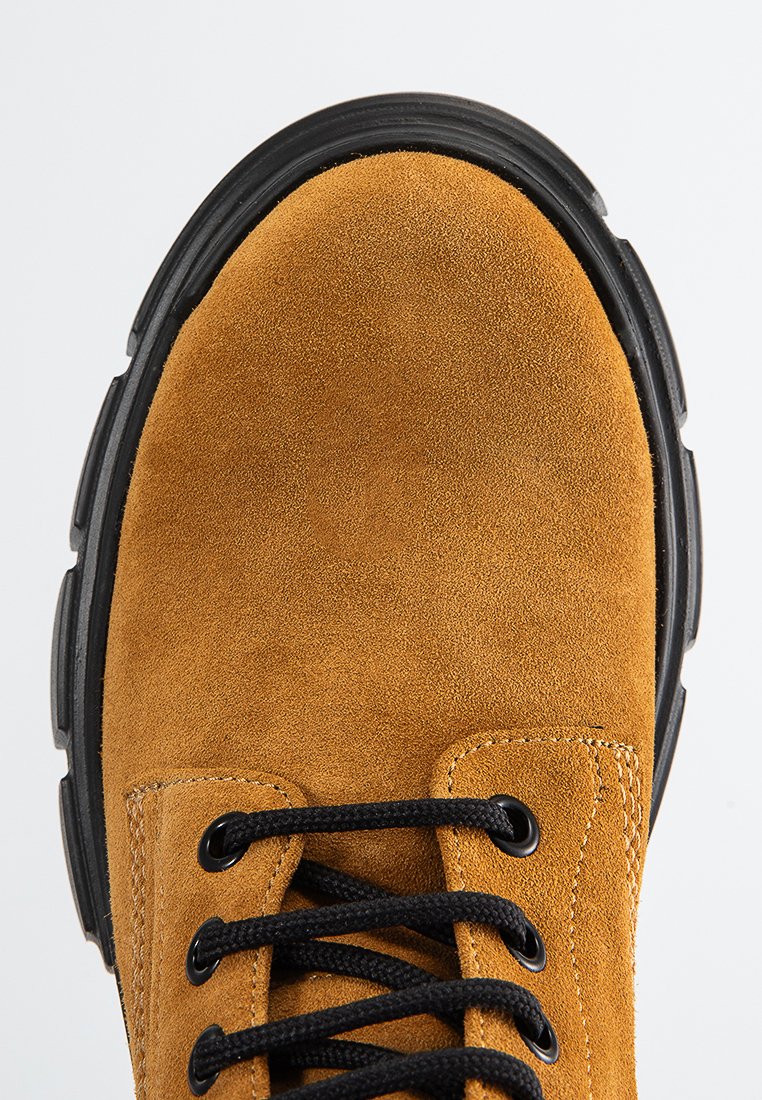 Bota de ante marrón con punta redonda, cordones negros y suela de goma. Presenta detalles cosidos y ojales para el cordón.