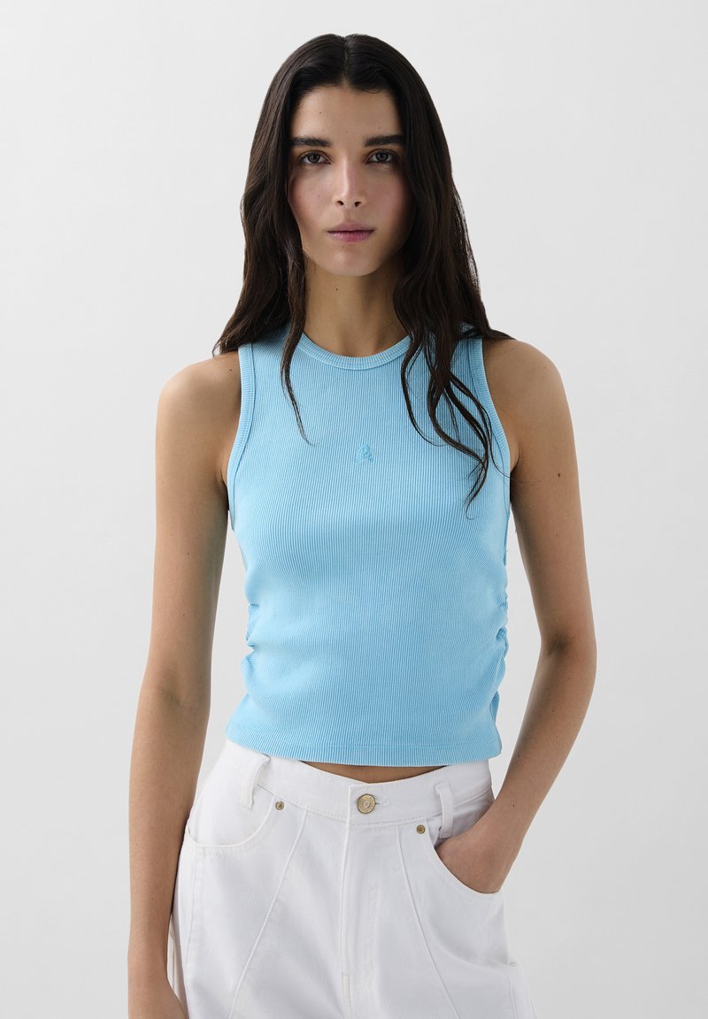 Lichtblauwe geribbelde tanktop met een ronde hals en plooien aan de zijkanten, gecombineerd met witte hoge taillebroek. Geen zichtbare logo's of patronen.