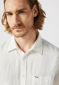 Biała lniana koszula z zapięciem na guziki, z małą kieszonką po lewej stronie na piersi oraz czarnym logo Wrangler. Gładka tekstura, standardowy kołnierzyk.