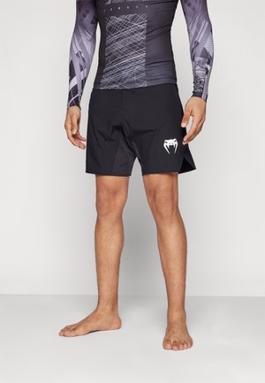 Man draagt zwarte sportshorts met wit slanglogo en een zwart-paarse sporttop met lange mouwen en patroon, staat op blote voeten.