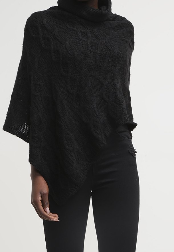 Personne portant un poncho en maille torsadée noire avec un col roulé et un pantalon noir ajusté sur un fond blanc uni.