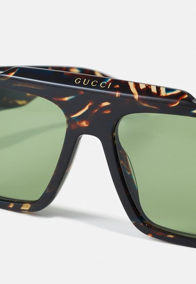 Gafas de sol Gucci con montura de carey oscuro, forma angular y lentes verdes. El logo dorado en la parte superior de la montura añade un detalle.