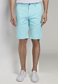 Uomo che indossa pantaloni corti azzurri fino al ginocchio e sneakers bianche, in piedi contro uno sfondo neutro.