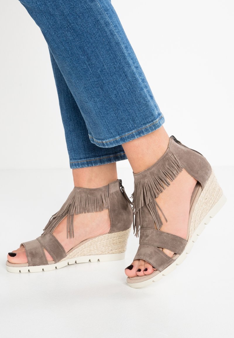 Pieds chaussés de sandales compensées en daim taupe avec des détails à franges et bouts ouverts, associées à un jean en denim bleu.