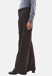 Bruine broek met wijde pijpen gemaakt van een gestructureerde stof met plooien aan de voorkant, gecombineerd met een grijs longsleeve topje en zwarte schoenen met hak.