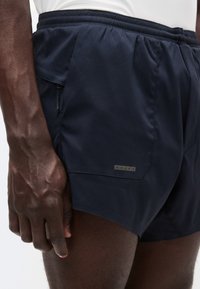 Shorts bleu marine avec une taille élastique, une poche latérale avec fermeture éclair, et une petite étiquette grise avec quatre trous près de la poche.