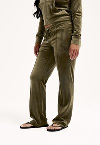 Zielony oliwkowy welurowy dres z krótką bluzą na suwak i spodniami z regulowanym paskiem w talii, wyposażonymi w kieszenie na zamek oraz teksturowane detale. Akrylowe klapki.