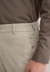 Homme portant un pantalon beige et une chemise à manches longues marron avec une texture, main détendue le long du corps, montrant les détails de la poche et de la ceinture.