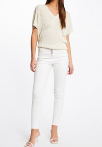 Kvinna som bär en krämfärgad kortärmad stickad topp med perforerade detaljer, högmidjade vita skinny jeans med knappar, samt vita klacksandaler.