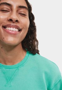 Felpa turchese con collo a giro, realizzata in cotone morbido. Presenta polsini e orlo a coste, con un focus su una vestibilità rilassata e un design minimal.